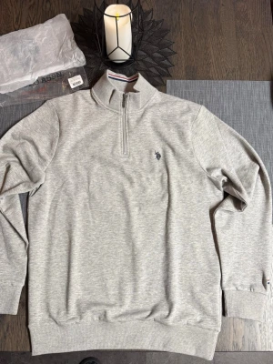 Grå half zip tröja från U.S. Polo Ralph Lauren - Snygg grå långärmad tröja från U.S. Polo Ralph Lauren med half zip och ståkrage med röd och blå randig detalj. Broderad logga på bröstet och ribbade muddar. Perfekt för lager-på-lager och chill stil.