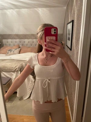 Vit topp med knyt från Nelly - Supersöt vit topp från Nelly med korta volangärmar och knyt i midjan. Toppens material är ribbat och den har en lätt utsvängd nederdel samt rundad halsringning. Perfekt för en fräsch och somrig look.