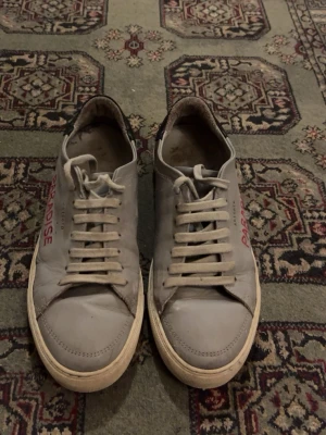 Axel Arigato  - Snygga vita sneakers från Axel Arigato med texten 'PARADISE' broderad i rött på sidan. Skorna har klassisk snörning, svart detalj vid hälen och är gjorda i skinn. Perfekta för dig som gillar stilrena men ändå unika skor. Säljer dom så här billigt för att vill bara bli av med dom så först till kvarn!