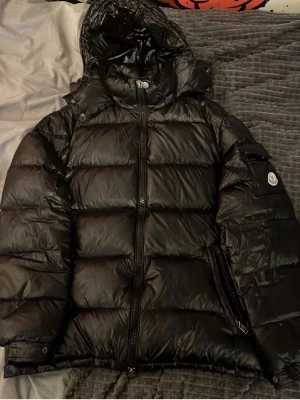 Svart Moncler pufferjacka med huva - Säljer en original Moncler Maya i svart. Jackan är använd och har viss slitning, vilket syns men påverkar inte funktionen nämnvärt. Den är fortfarande varm, bekväm och perfekt för vinterbruk.  Det finns ett mindre problem med dragkedjan längst ner, men i övrigt fungerar jackan som den ska. Perfekt för dig som vill ha en klassisk Moncler till ett bättre pris.  Hör av dig vid intresse eller frågor!  Vill du ha en kortare version, eller en som 