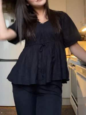 Svart blus med knyt och v-ringning - Säljer en svart blus med v-ringad hals och korta ärmar. Toppen har en avslappnad passform med knyt i midjan som ger en fin siluett. Tillverkad i ett lätt och luftigt material, perfekt för dig som gillar en enkel men snygg look.