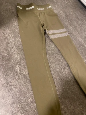 Olivgröna leggings från Stronger - Snygga olivgröna leggings från Stronger i storlek XS. Hög midja med logga runt hela linningen och två vita randiga detaljer på ena benet. Tillverkade i stretchigt material av 75% återvunnen polyester och 25% elastan. Perfekta för träning eller chill.