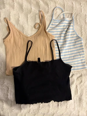3-pack croppade linnen i olika färger - Säljer tre croppade linnen: ett ribbat beige med v-ringning ett svart med smala axelband och vågig nederkant som är från H&M samt ett blåvitt randigt med raka axelband som är ifrån Bikbok Perfekta för sommaren och lätta att matcha till jeans eller kjol. Alla är I storlek XS