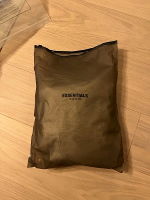 Essentials Fear of God mjukisbyxor beige - Snygga mjukisbyxor från Essentials Fear of God i en ljusbeige färg. Byxorna har elastisk midja med snörning och logga tryckt på benet. Perfekta för en chill och avslappnad stil. Tillverkade i mjukt bomullsmaterial för extra komfort.