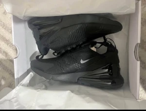 Nike Air Max 270 svarta sneakers - Nike Air Max 270 sneakers i helsvart färg med en stor synlig Air-enhet i hälen. Ovandelen är i mesh och syntet, med snörning och en liten silverfärgad swoosh på sidan. Skorna har en rund tå och platt sula, perfekt för dig som gillar sportig stil.