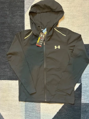 Svart vindjacka från Under Armour - Svart vindjacka från Under Armour med huva och hel dragkedja. Jackan har diskreta gula detaljer och logga på bröstet samt stort Under Armour-tryck på ryggen. Tillverkad i lätt syntetmaterial, perfekt för träning utomhus. Storm-teknologi för extra väderskydd.