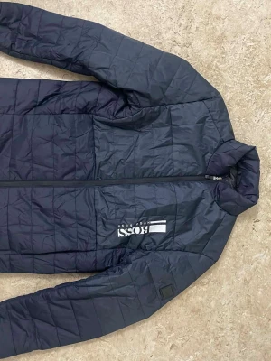 Mörkblå quiltad pufferjacka från BOSS - Säljer en mörkblå quiltad pufferjacka från BOSS med diskret logga på bröstet. Jackan har lång ärm, dragkedja framtill och en stilren, sportig look. Perfekt för kyligare dagar och enkel att matcha med olika outfits.