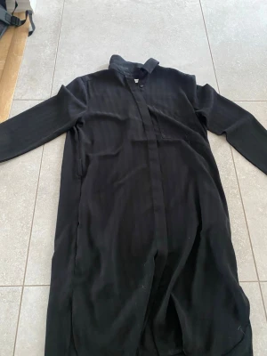 Svart långklänning Carin Wester - Säljer en svart långklänning från Carin Wester i storlek 36. Klänningen har diskreta ränder, lång ärm och klassisk krage. Stängs med knappar framtill och har en stilren, minimalistisk look. Perfekt för dig som gillar enkel men snygg design.