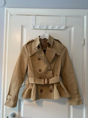 Kort kappa / trenchcoat - Slutpris!!! Helt ny jacka med prislapp kvar. Klassisk trench-inspirerad modell i beige med skärp i midjan och dubbla knapprader. Väldigt stilren och tidlös design som påminner om B**berry, dock INTE från det märket. Bandet går att justera och ta av och på. Alla knappar går att knäppa av och på. Volang aktigt längst nere   Storlek står som 36, men jag skulle snarare säga att den passar 34 bäst. Högkvalitativ känsla och fint fall.  