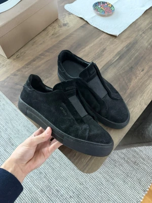 Svarta sneakers från Arigato i mocka - Säljer ett par svarta sneakers från Arigato i mjuk mocka med minimalistisk design. Skorna har elastisk panel framtill istället för snörning och en tjock, svart platt sula. Perfekta för dig som gillar stilrena och bekväma skor.