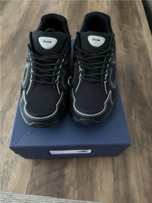 CD b30 - Säljer ett par svarta Di0r sneakers med vita kontrastdetaljer och snörning. Skorna har en chunky sula och meshpaneler för extra komfort. Märket syns tydligt på plösen och sidan. Perfekta för dig som vill ha en clean och modern look.