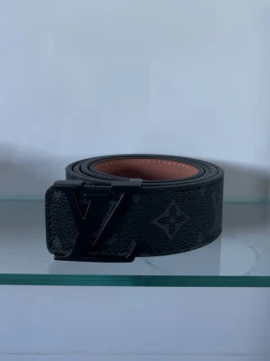 Svart monogram skärp - Säljer ett svart skärp med klassiskt monogrammönster och stor logga på spännet. Bältet är gjort i präglat skinn med brun insida och bred design som sticker ut. Perfekt accessoar för att lyfta vilken outfit som helst.