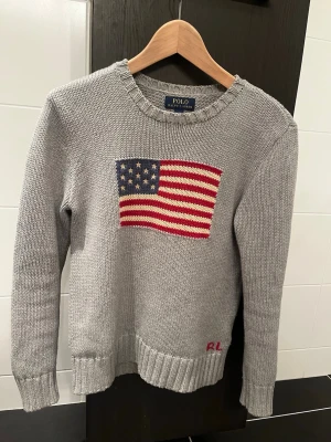 Grå stickad tröja Polo Ralph Lauren - Snygg grå stickad tröja från Polo Ralph Lauren med amerikansk flagga framtill. Tröjan har rund hals, ribbade muddar och RL-broderi nertill. Perfekt för dig som gillar klassisk och avslappnad stil.