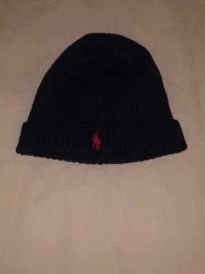 Mörkblå mössa från Polo Ralph Lauren - Säljer en mörkblå ribbstickad mössa från Polo Ralph Lauren med klassisk röd broderad logga framtill. Mössan är i mjukt material och har vikt kant för extra stilpoäng. Perfekt accessoar för kyliga dagar.