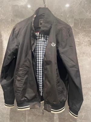 Svart bomberjacka från Etirel - Säljer en svart bomberjacka från Etirel Campus Sportswear i storlek small. Jackan har vita ränder på muddar och krage, dragkedja framtill och ett rutigt innerfoder i blått och vitt. Klassisk college-look med broderad logga på bröstet.