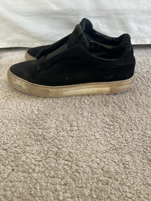 Svarta slip-on sneakers från Axel Arigato - Svarta slip-on sneakers från Axel Arigato i mocka med vit, något smutsad gummisula. Klassisk låg modell utan snörning och med diskret logga på plösen. Skorna har en enkel och clean design som passar till det mesta.