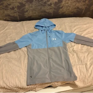 Blå och grå vindjacka från Under Armour - Säljer en snygg tvåfärgad vindjacka från Under Armour i blått och grått. Jackan har huva, dragkedja framtill och två sidofickor. Perfekt för dig som vill ha en lätt och sportig jacka med stilren design och logga på bröstet.