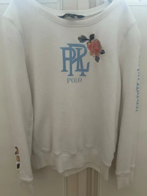 Vit sweatshirt Polo Ralph Lauren blommig - Vit sweatshirt från Polo Ralph Lauren med ljusblå logga på bröstet och broderade blommor i rosa och beige på bröstet och ena ärmen. Tröjan har rund halsringning och långa ärmar. Skön och stilren med snygga detaljer.