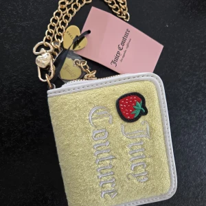Juicy Couture plånbok med jordgubbsmärke - Superfin plånbok från Juicy Couture i ljusgul sammet med broderad logga och en söt jordgubbsdetalj. Baksidan har fack med fönster för kort, och insidan är fodrad i rosa med tryckt logga. Kedja i guld med hjärtberlocker ingår.