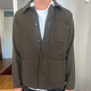 Grön overshirt från Asket i ull - Stilren grön overshirt från Asket med tre stora fickor framtill och klassisk krage. Jackan är tillverkad i mjuk ull och har en rak, normal passform med knäppning framtill. Perfekt för lager-på-lager och enkel att matcha med olika outfits. Perfekt till hösten! 