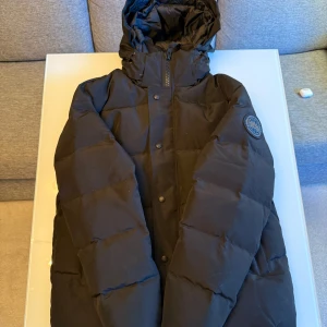Svart dunjacka från Canada Goose M - Säljer en svart dunjacka från Canada Goose i storlek M. Jackan har huva, dragkedja och knappar framtill samt logotyp på ärmen. Fodrad insida med meshfickor och klassisk pufferstil som håller dig varm under kalla dagar.