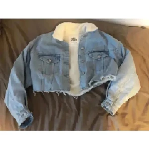 Zara Croppad jeansjacka med teddyfoder - Snygg Zara croppad jeansjacka i ljusblå denim med vitt teddyfoder på insidan och kragen. Jackan har två bröstfickor med knappar, rå kant nertill och långa ärmar. Perfekt för dig som vill ha en cool och trendig look.