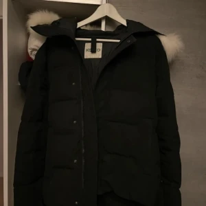 Svart pufferjacka från Kenzo med fuskpäls - Kenzo pufferjacka i svart med stor vit fuskpäls på huvan. Jackan har dragkedja och tryckknappar framtill, samt diskret Kenzo-logga på ärmen. Perfekt för kalla dagar med sin vadderade design och snygga detaljer.