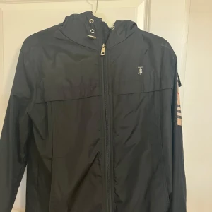 Svart Burberry vindjacka med rutig detalj - Svart vindjacka från Burberry med dragkedja framtill, huva med snörning och klassiskt rutigt foder i beige, svart och rött. Ärmen har en patch med Burberry-logga och rutigt tyg. Jackan har en clean och sportig look, perfekt för dig som gillar stilrena detaljer.