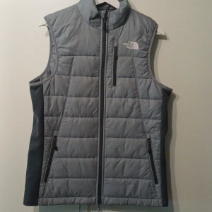 Grå dunväst från The North Face - Snygg grå dunväst från The North Face med quiltad design och hög krage. Västen har tre dragkedjeförsedda fickor framtill och broderad logga på bröstet. Perfekt för lager-på-lager och outdoor-vibes.