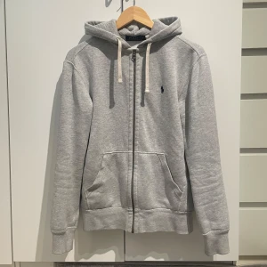Grå hoodie från Polo Ralph Lauren - Grå hoodie från Polo Ralph Lauren med dragkedja, huva med vita snören och klassisk blå logga på bröstet. Mjuk bomullskänsla, känguruficka framtill och ribbade muddar. Perfekt för chill dagar och enkel att matcha med jeans eller joggers.