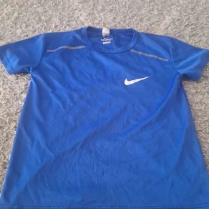 Blå Nike t-shirt med logga - Snygg blå t-shirt från Nike med klassisk vit swoosh-logga på bröstet. Tillverkad i mjuk polyester som andas, perfekt för träning. T-shirten har rund hals och korta ärmar, samt diskreta grå detaljer vid axlarna. Enkel och sportig stil.