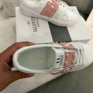 Valentino Rockstud vita sneakers - Snygga vita Valentino Rockstud sneakers i skinn med ljusrosa band och ikoniska nitar över vristen. Klassisk rund tå, platt sula och snörning framtill. Märkeslogga på plösen och sula med mönstrad struktur. Perfekta för dig som gillar exklusiv streetstyle.