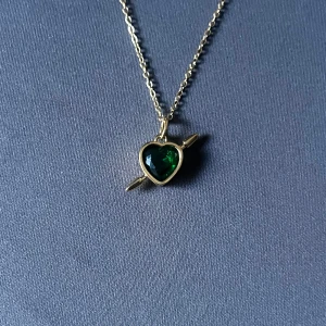 Rebel Heart – Golden Emerald -  Guldpläterat halsband i rost- och nickelfritt stål. Smaragdgrönt hjärta med pil. Kedja 50 cm, justerbar. Hängets diameter 0,9 cm. 