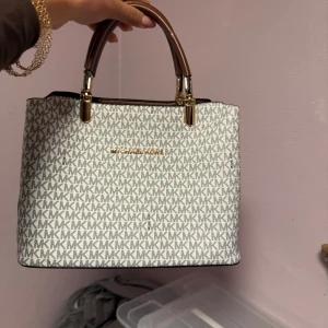 Väska - Snygg handväska från Kors med klassiskt -mönster i grått och vitt. Väskan har bruna handtag med guldfärgade detaljer och en fyrkantig form. Perfekt storlek för att få plats med allt viktigt och ger en lyxig vibe till din outfit.