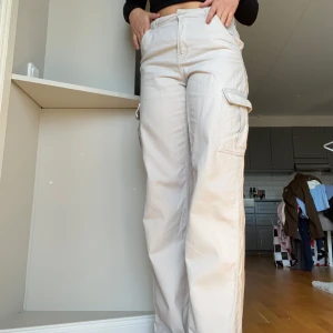 Beiga cargopants med raka ben - Säljer ett par beiga cargopants med raka ben och stora sidofickor. Byxorna har hög midja och är tillverkade i bomull.  jag har sytt ner dom men sena ena sidan har släppt en aning ( se sista bilden)