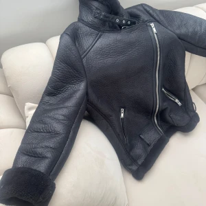 Svart pälsjacka från Zara Woman XS - Säljer en svart pälsjacka från Zara Woman i storlek XS. Jackan har en cool bikerstil med sned dragkedja, högt krage med spänne och mjukt fuskpälsfoder. Utsidan är i skinnimitation och jackan har flera fickor med dragkedja samt uppvikta ärmslut.