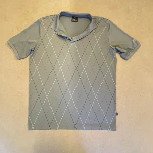 Grå mönstrad piké från Oscar Jacobson - Snygg grå pikétröja från Oscar Jacobson med klassiskt rutmönster i vitt, svart och rött. Tröjan har korta ärmar, krage och knäppning framtill. Perfekt för en stilren look och passar till många tillfällen.