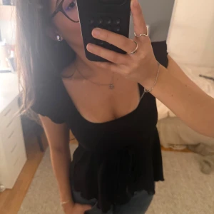 Svart blus med knyt i midjan - Säljer en svart blus med korta ärmar och fyrkantig urringning. Toppen har ett knytband i midjan som ger en snygg siluett och lätt volang nertill. Perfekt att styla med jeans eller kjol för en trendig look.