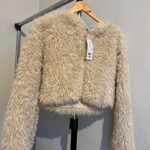 Beige pälsjacka från Gina - Säljer en fluffig och croppad beige pälsjacka från Gina tricot storlek XS. Jackan har lång ärm, rund halsringning och är gjord i mjukt syntetmaterial som ger en riktigt mysig look.