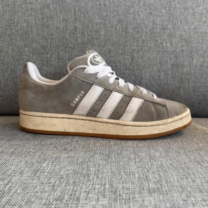 Adidas Campus grå sneakers mocka - Säljer ett par klassiska Adidas Campus sneakers i grå mocka med vita detaljer och gummisula. Skorna har de ikoniska tre ränderna på sidan, snörning och låg profil. Perfekta för dig som gillar retrostil och vill ha något som funkar till det mesta. Nypris: 1500kr✅💯