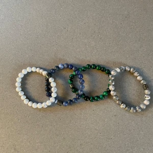 Fyra pärlarmband i olika färger - Säljer ett set med fyra elastiska armband av runda pärlor i olika färger: vitt, blått, grönt och gråmelerat. Varje armband har små metalliska detaljer som ger en cool touch. Perfekt för att mixa och matcha till din stil.