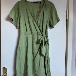 Grön omlottklänning med knyt - Snygg oanvänd grön omlottklänning från Veromoda med korta ärmar och knyt i midjan. Klänningen har v-ringning. Perfekt för varma dagar.