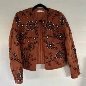 Broderad brun quiltad jacka från MANGO - Superfin brun quiltad jacka från MANGO med broderade blommor i svart och rött samt paljettdetaljer. Jackan har rund halsringning, lång ärm och stängs framtill. Perfekt för dig som vill sticka ut med en unik och bohemisk stil.🤩💕