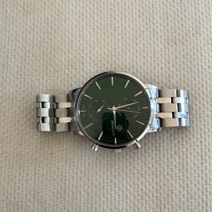 GANT klocka med grön urtavla - Stilren klocka från GANT med mörkgrön urtavla och silverfärgat armband i rostfritt stål. Urtavlan har minimalistiska index, två subdials och snygga detaljer. Länken är robust och har GANT-logga på spännet. Perfekt accessoar för dig som gillar klassisk design.