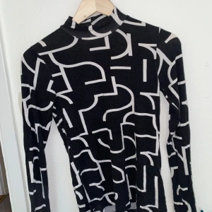 Svartvit polotopp med grafiskt mönster - Säljer en svart polotopp med vita grafiska linjer. Toppen har lång ärm och är figurnära i modellen. Perfekt att styla med jeans eller kjol för en cool look.