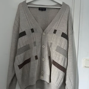 Beige kofta från Greenfield med knappar - Snygg beige kofta från Greenfield med v-ringning och stora knappar framtill. Koftan har ett diskret mönster med svarta och bruna ränder samt ribbade muddar. 