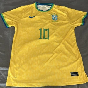 Brasilien Ronaldinho 10 Nike fotbollströja - Gul Brasilien fotbollströja från Nike i storlek L med gröna och blå detaljer, nummer 10 och Ronaldinho på ryggen. Broderat CBF-märke på bröstet och blå Nike-logga. Tillverkad i lätt Dri-FIT-material, perfekt för match eller träning. Tröjan är i nyskick.