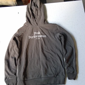 Svart hoodie från Peak Performance - Svart hoodie från Peak Performance med vit logga på bröstet. Klassisk modell med huva och känguruficka framtill. Tillverkad i mjuk bomullsmix, perfekt för chill dagar eller när du vill ha en enkel och snygg look.