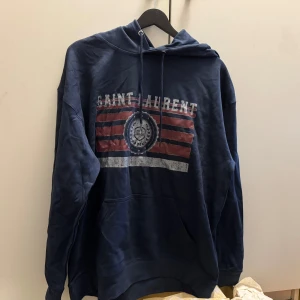 Blå Saint Laurent hoodie med tryck - Säljer en marinblå hoodie från Saint Laurent med stort tryck framtill i rött, vitt och blått. Tröjan har huva med snörning och en stor magficka. Perfekt för dig som gillar streetwear och vill ha en snygg och bekväm look.