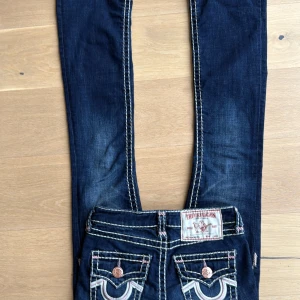 True Religion Becca Bootcut Jeans - Mörkblå limited edition jeans från True Religion, modell Becca med bootcut. Snygga kontrastsömmar i vitt och rosa, klassiska fickor med dekorativa detaljer och kopparfärgade knappar. Midwaist/lowwaist. Ny pris 2700 men säljer för 2000 så dom är limited edition och knappt använda.